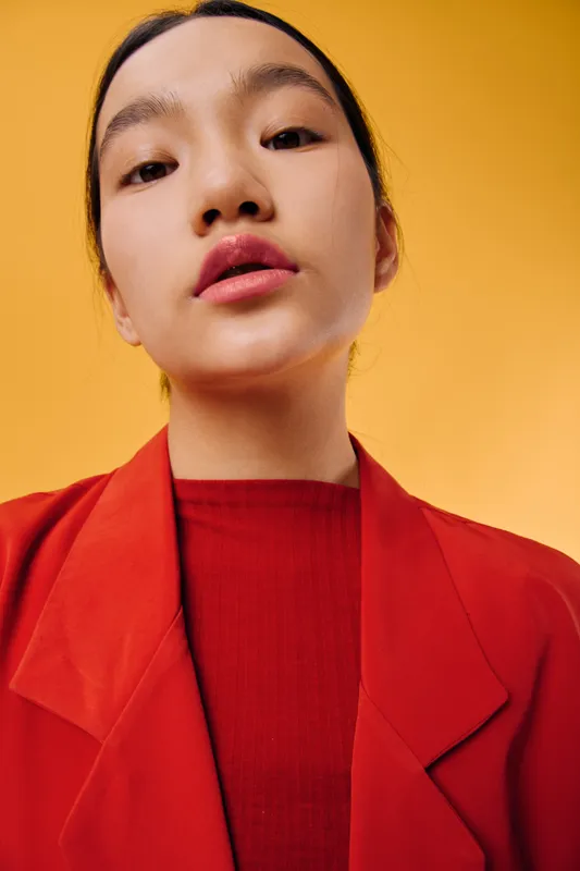 Young woman wearing red coat and sweater looking confidently into the camera with neutral expression on yellow background. Portrait, fashion, beauty, and modern style in studio.