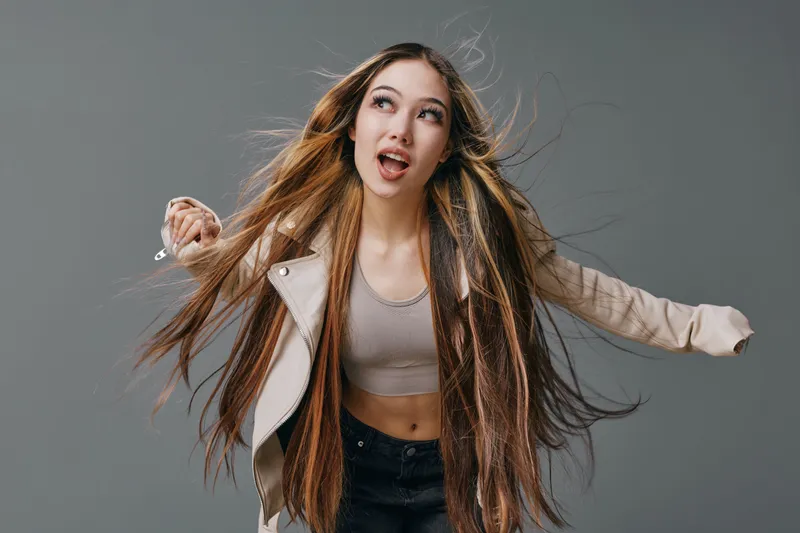Young woman with long braided hair holding makeup brush and looking up, wearing casual jacket and crop top, posing on grey background in studio.
