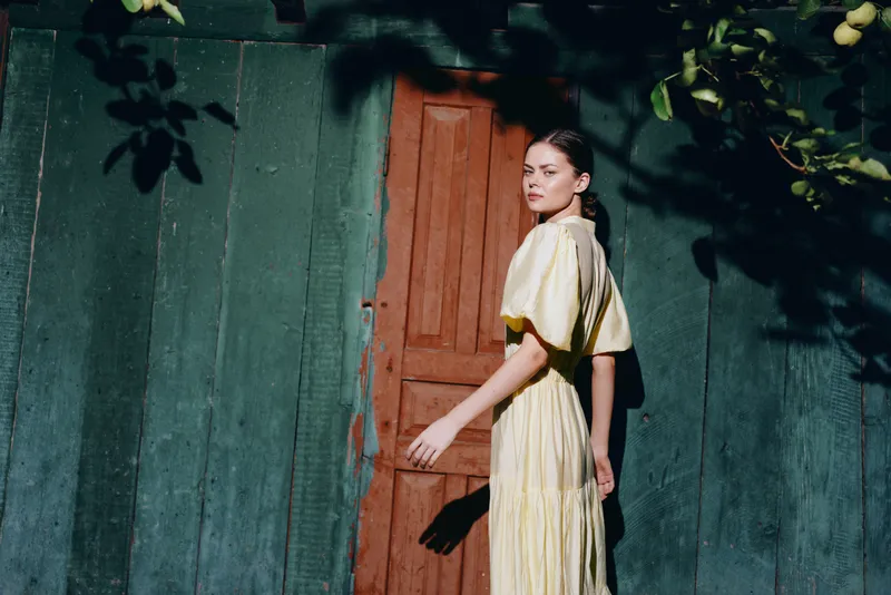 Woman in yellow dress by wooden door, sunlight and shadow pattern on wall, candid portrait conveying authenticity inclusivity and calm
