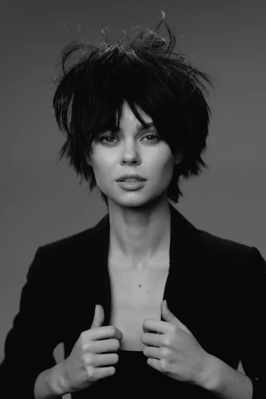 Black and white portrait of a stylish woman with tousled hair and intense gaze wearing a dark jacket.