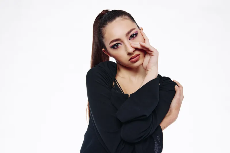 Young stylish woman with bold pink eye makeup and sleek high ponytail poses with one hand on her face and the other hugging her shoulder against a white background. Beauty fashion concept.