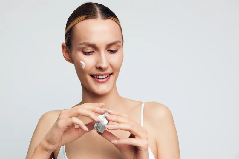Young woman applies moisturizing cream on face holds open jar smiles with natural skin care routine isolated on white background beauty health concept closeup portrait.