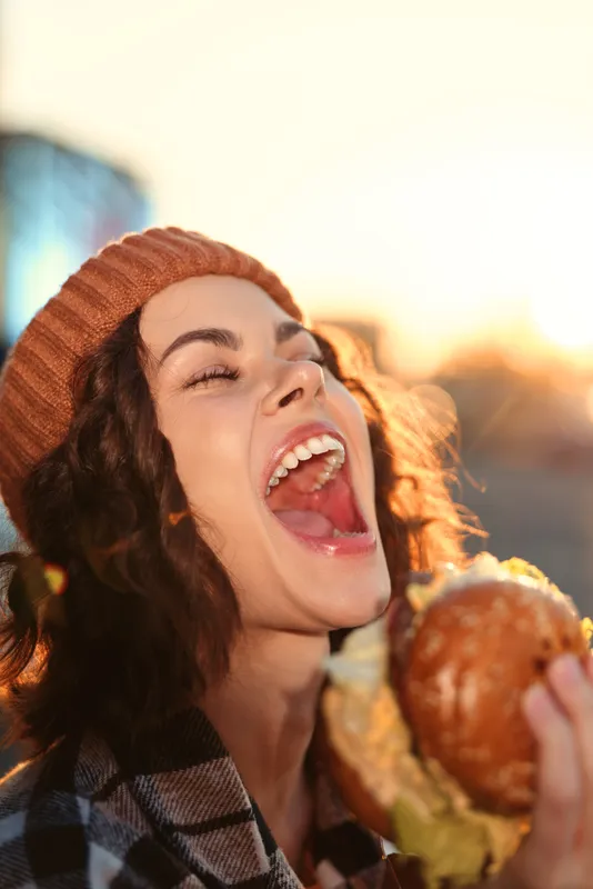 Woman smiling and laughing while biting a burger at golden hour, candid urban moment with authenticity and candid lifestyle vibe, warm golden hour glow and emotional storytelling.