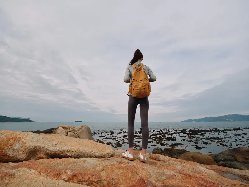 A young woman stands on rocky terrain by the sea, wearing a yellow backpack, grey leggings, and a denim jacket, conveying a sense of adventure and exploration