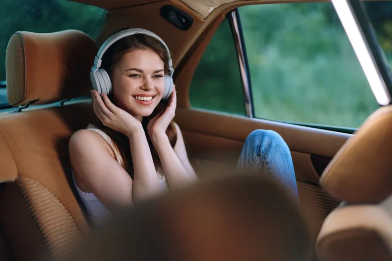 Young woman enjoying music in a car, wearing headphones and smiling, showcasing relaxation and happiness in a natural setting.