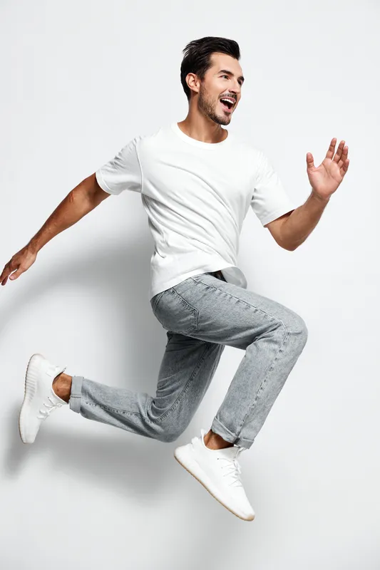 Energetic smiling man wearing white t-shirt jumps joyfully against a plain light background. Captivating moment showcasing enthusiasm and happiness.