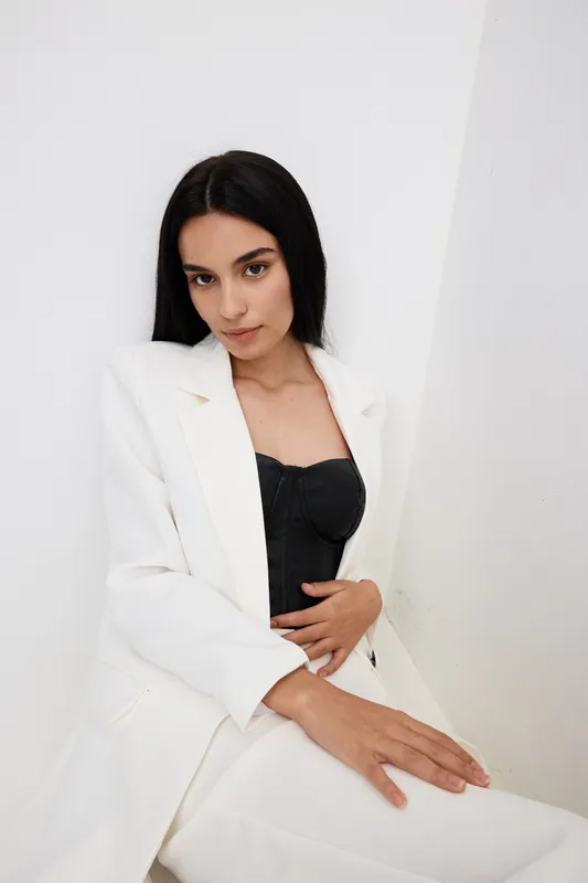 young woman in white suit and black top seated indoors with thoughtful expression against minimalist white wall, modern fashion portrait with natural lighting