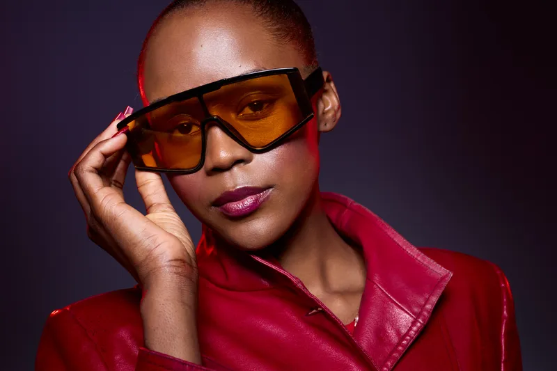 Elegant African woman with dark skin wears oversized amber sunglasses and a red leather jacket. Studio portrait with dramatic lighting highlights fashion and confident style.