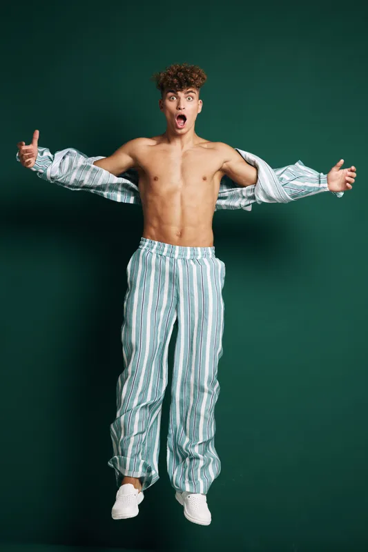 Young man jumping with open shirt and striped pants on green background, showing surprised expression and casual style with white sneakers in studio photo shoot.