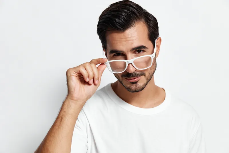 Charming man wearing glasses smiles confidently at the camera while adjusting his eyewear, showcasing a modern and stylish look against a light background.