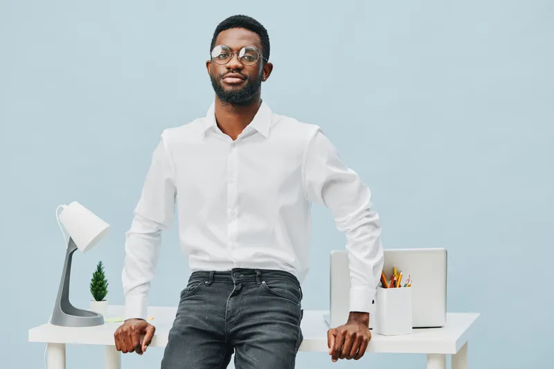 Confident young African American man in white shirt stands at modern desk with laptop, lamp, and stationery on light blue background, studio shot. People lifestyle concept