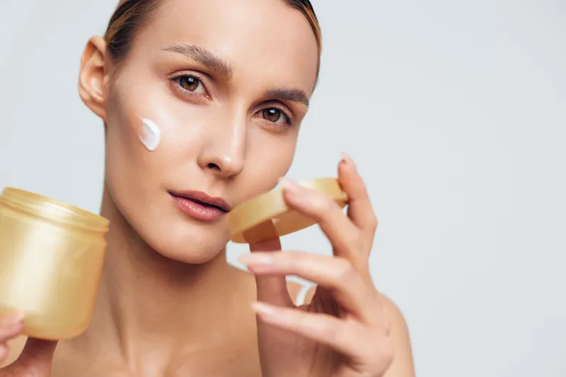 Woman applying face cream from a small jar on her cheek and under eye in a bright studio, calm beauty routine, skincare hydration and cosmetic care daily concept