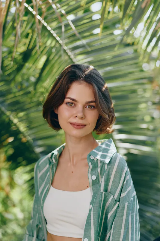 Portrait of a young woman standing outdoors among lush palm leaves, wearing a striped green shirt over a white crop top.