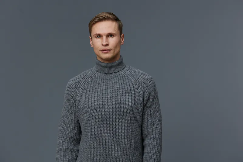 Modern portrait of a handsome young man with fair hair and blue eyes, wearing a cozy gray knitted sweater, looking confidently at the camera on a plain gray background. Studio shot for fashion and lif