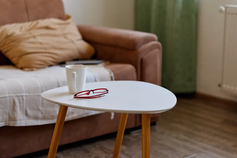 Cozy living room scene with a white round table in the foreground, a cup resting on it, a sofa and soft pillows in the background, conveying a calm intimate home moment