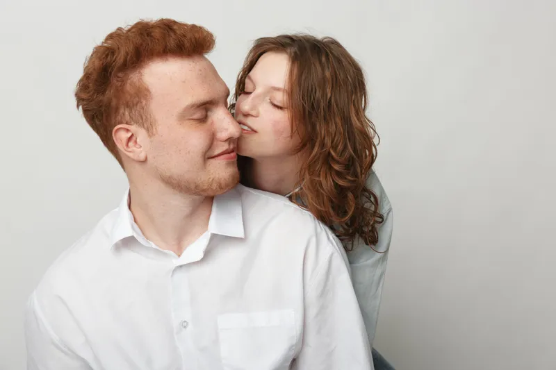 Young couple in casual clothes share intimate moment, woman gently kisses man on cheek. Light background creates soft romantic atmosphere, concept of love and affection between partners.
