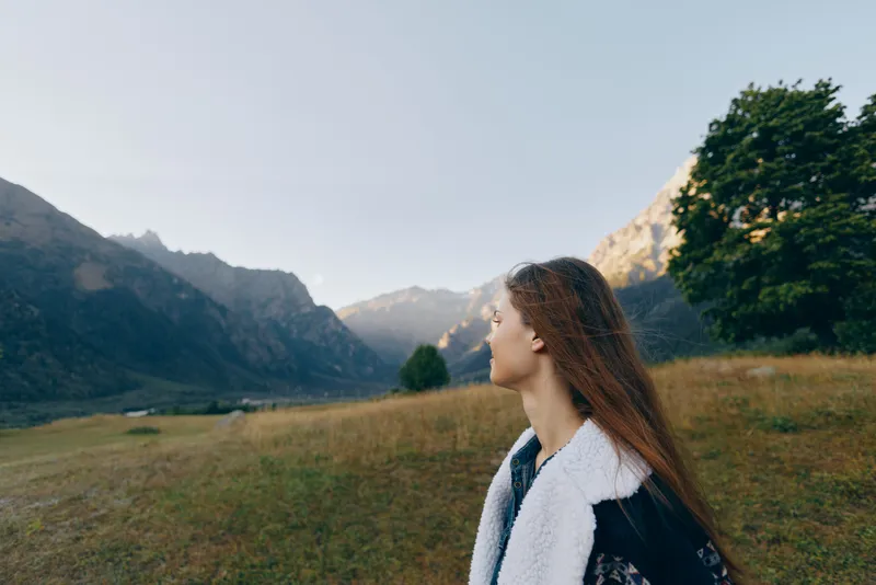 Woman in profile looks at distant mountains across a wide meadow landscape, a nature scene outdoors and travel concept with peaceful mood, long hair and layered jacket.