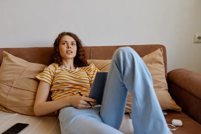 A young woman relaxes on a sofa at home with a tablet, wearing a striped yellow t shirt and light blue jeans, looking engaged, comfortable, and focused in a cozy living room with soft daylight.