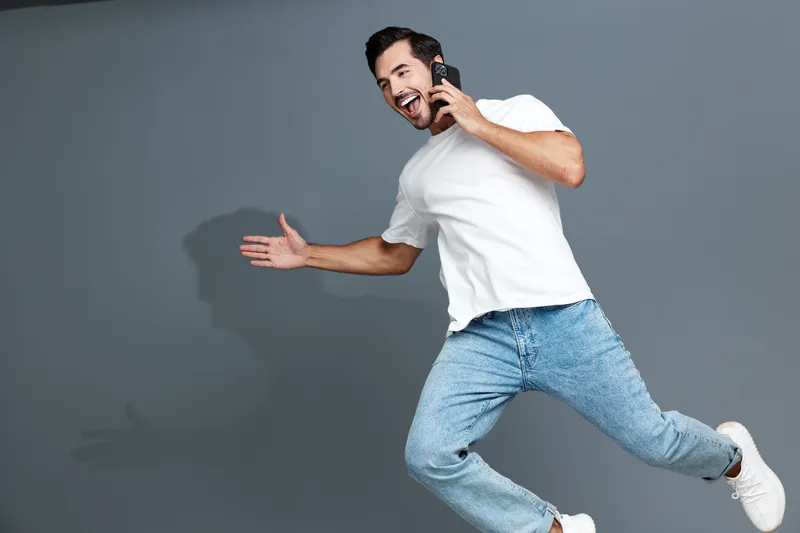 Full body smiling cheerful young man wear casual white t-shirt look camera talking on mobile phone isolated on plain solid gray background studio. People lifestyle concept
