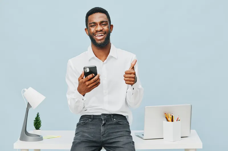 Smiling man holding smartphone, giving thumbs up, standing in modern office with desk, laptop, stationery, and minimal decor, cheerful mood, professional lifestyle concept, business casual attire, nat