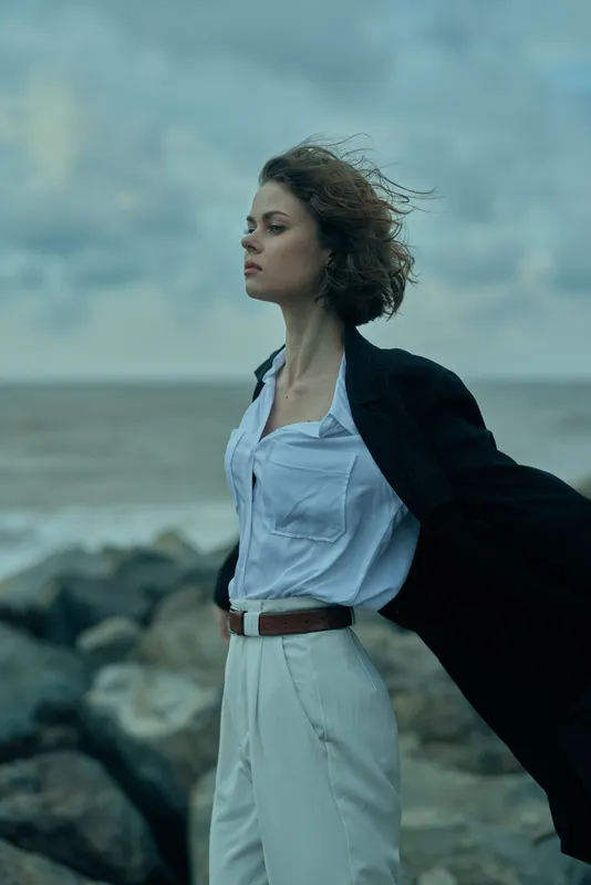 Wind swept woman on a windy coast wearing a light blue shirt and white trousers, blazer open, hair in motion, conveys confidence and freedom.