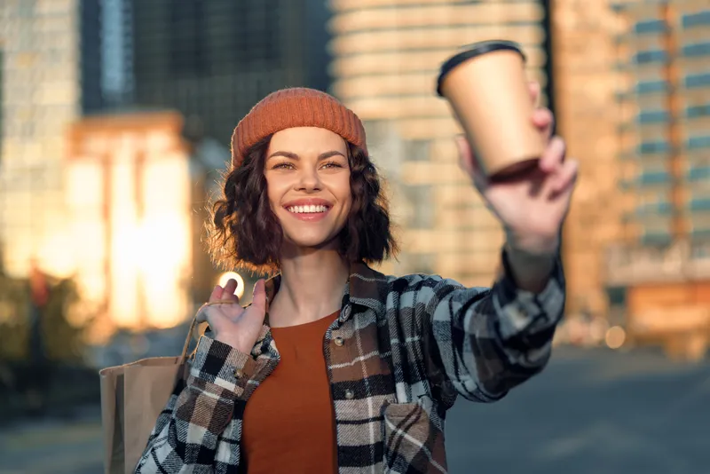 woman coffee smile urban beanie candid authenticity golden glow. Candid lifestyle portrait of a joyful millennial holding takeaway cup at golden hour, mindful living and emotional storytelling.
