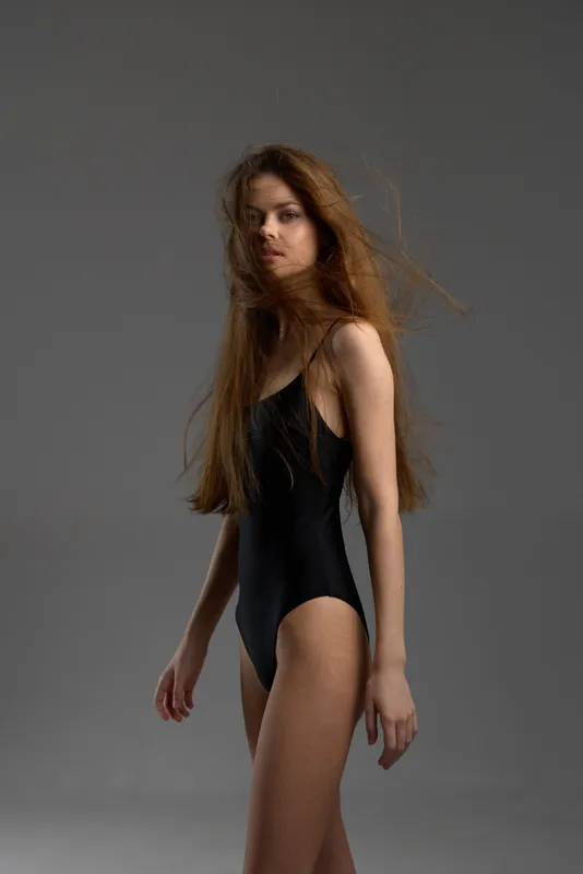Young woman in black swimsuit posing against neutral gray background with flowing hair and calm expression