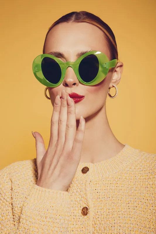 Stylish woman with green round sunglasses and red lipstick covers mouth with hand. She wears yellow knitted sweater and gold hoop earrings on yellow background. Fashion and beauty concept.