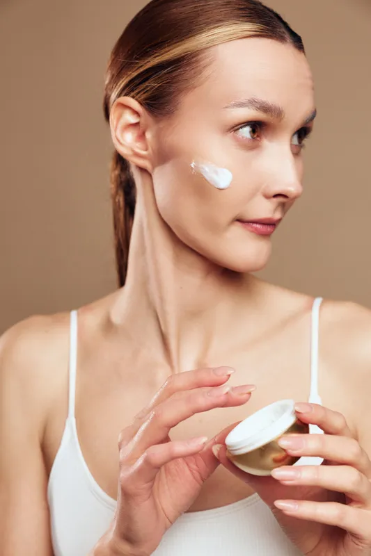 Young woman applies moisturizing cream on her cheek skincare beauty routine natural healthy skin care face closeup isolated on beige background in white top.