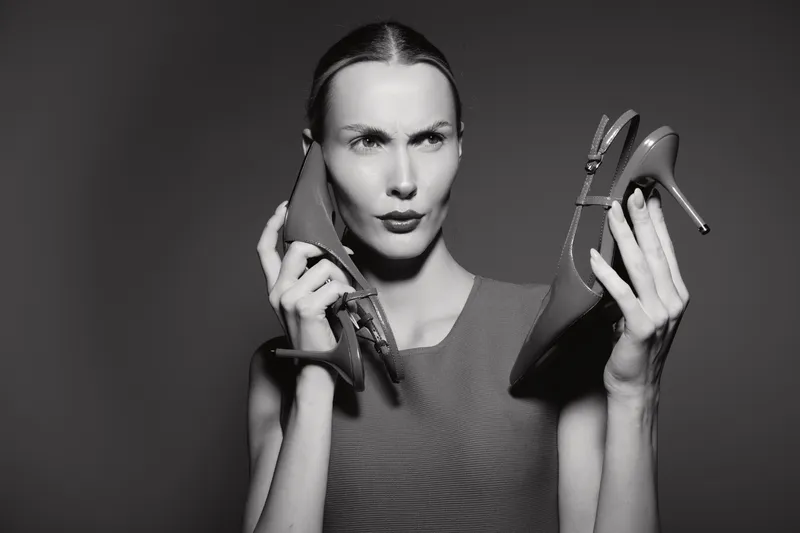 A young Caucasian woman with fair skin holds high heel shoes near her face, expressing confusion or frustration. Concept of fashion dilemma, choice, and footwear decision making.