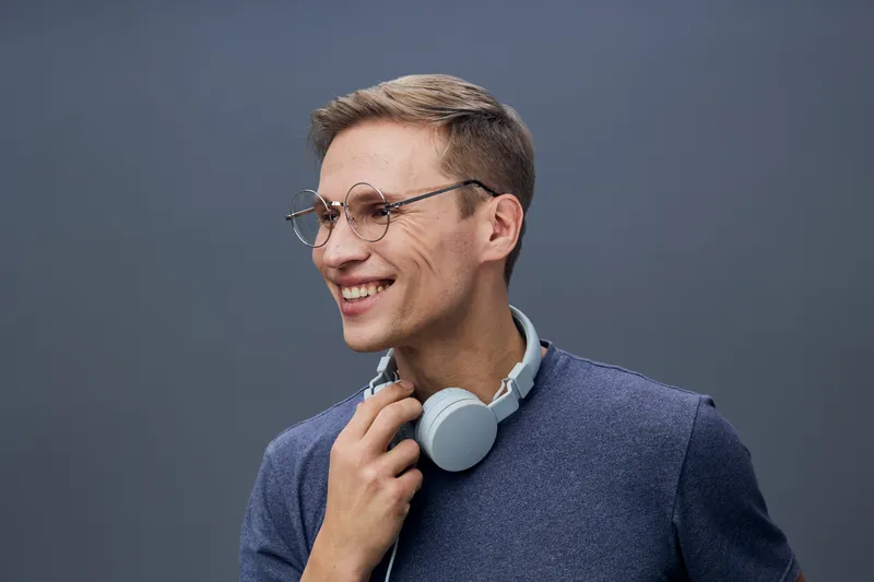 Smiling young man with glasses and headphones around neck, casual style, modern appearance, cheerful expression, trendy fashion, indoor studio portrait, gray background, lifestyle concept, happy mood