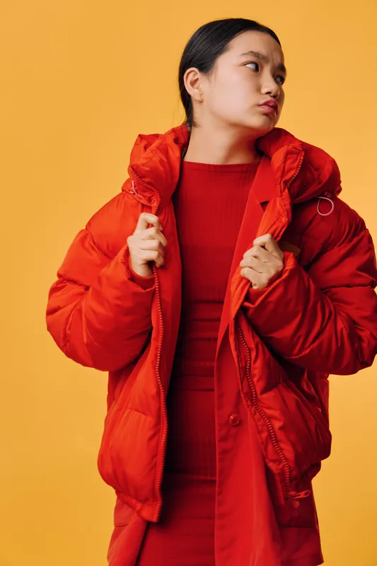 Woman in red puffer jacket and matching dress standing against yellow background holding jacket collar, looking to the side with confident expression in studio portrait fashion clothing style.