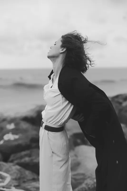 Windy beach scene with a woman in light trousers and white shirt, a long coat billowing behind, rocky shore and sea in the distance, dramatic motion and freedom