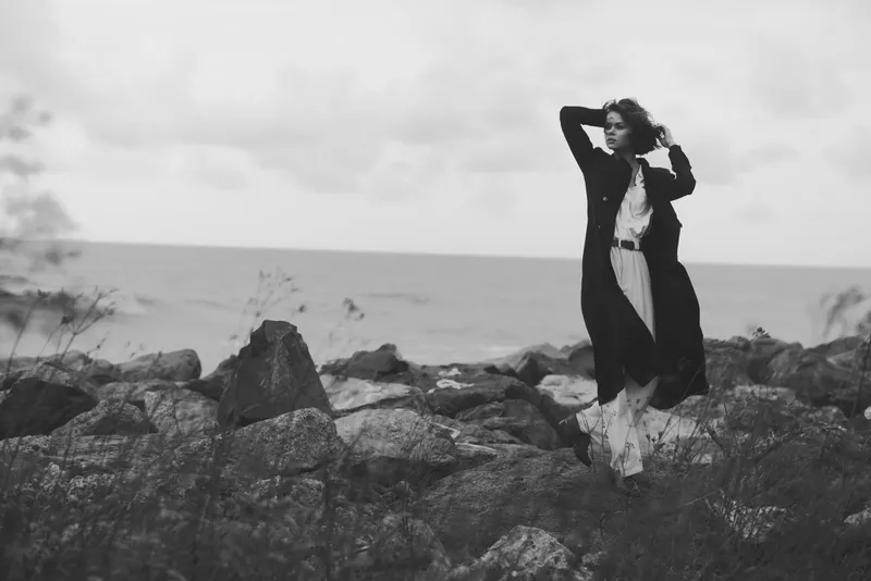 Fashionable woman wearing a long coat on a rocky shore, wind blowing through her hair, standing by the sea to create a dramatic outdoor landscape