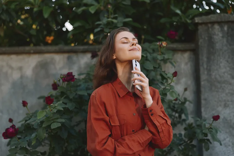 Woman lifestyle outdoor portrait among garden roses, wearing a rust red blouse, smiling softly with eyes closed, enjoying a moment of selfcare and fragrance in a peaceful setting