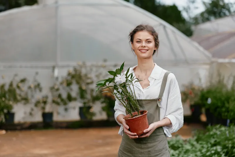 Young woman holding a potted plant in a greenhouse, wearing casual overalls, showcasing a joyful expression, surrounded by lush greenery and soft natural lighting