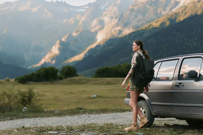 Young woman with backpack standing next to car in mountain landscape. Outdoor adventure, travel lifestyle, hiking concept with natural sunlight and scenic background.