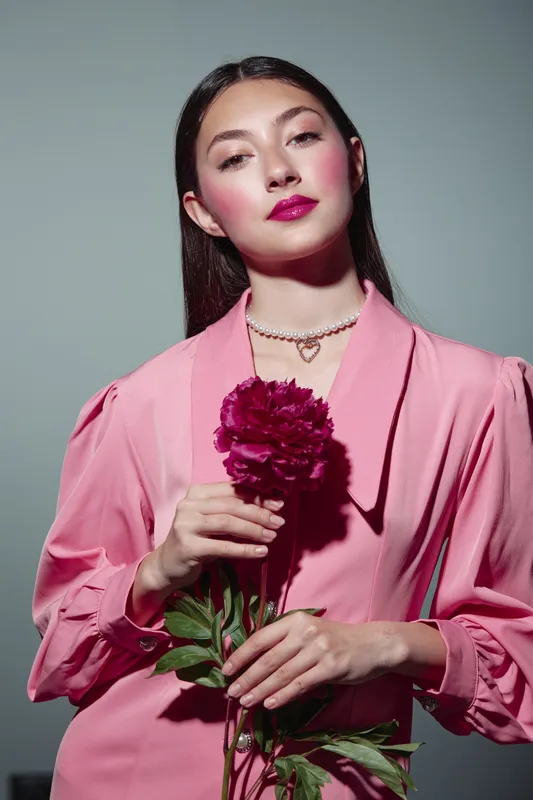 Confident young woman in pink blouse holds dark red peony while posing against neutral background. Concept of fashion, beauty, elegance, and creative portrait photography.
