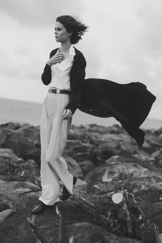 Woman stands on rocky shore, wind-swept cape behind her, wearing shirt and blazer, monochrome outdoor fashion shot with ocean backdrop and strong, dramatic stance.