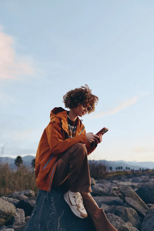 Woman lifestyle outdoors at sunset, curly woman in orange jacket sitting on rocks, looking at smartphone, relaxed posture, warm natural light, soft sky, calm smile