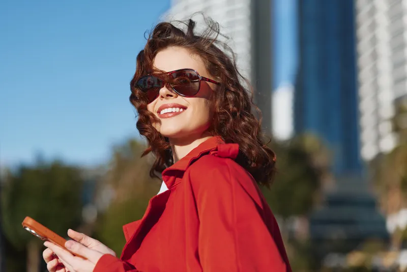 Young woman with curly hair wearing a red coat and sunglasses smiles while holding a smartphone outdoors in an urban setting on a sunny day.