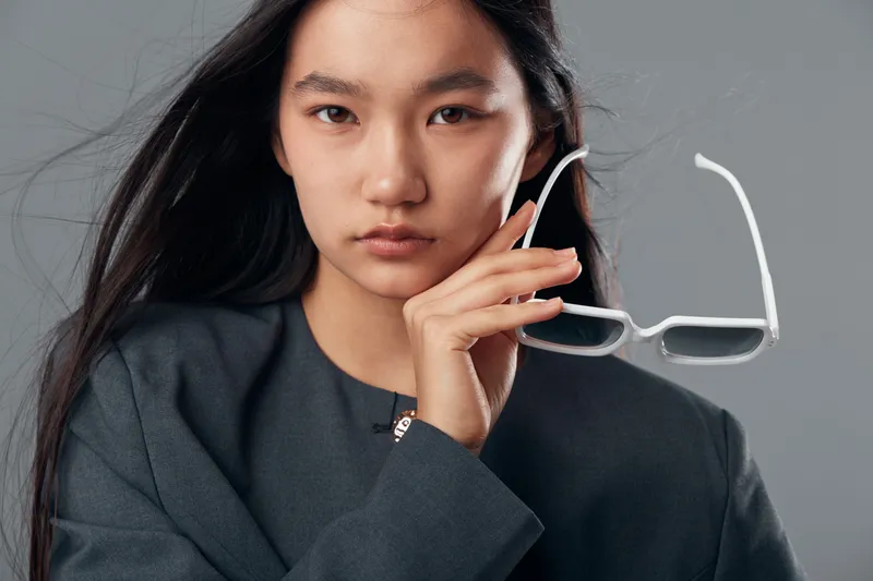 Young woman with long black hair holding transparent eyeglasses close to her face wearing dark clothing against neutral background with serious expression.