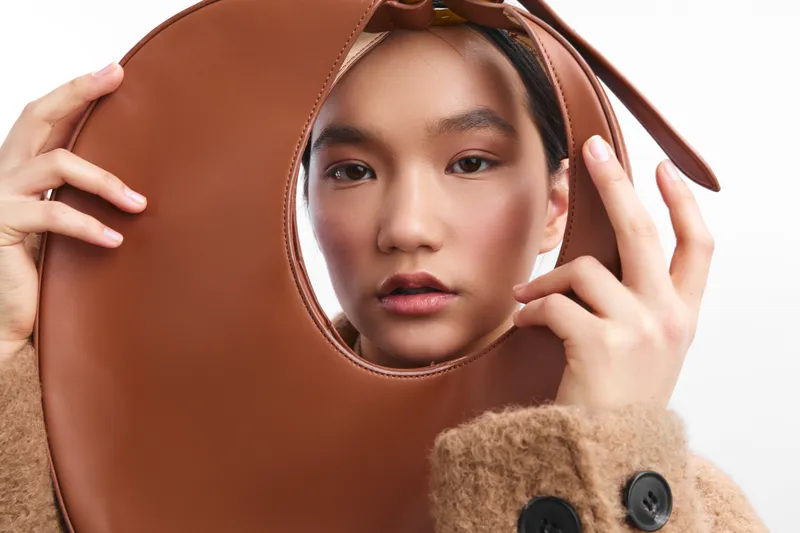 Asian woman gazes softly at the camera while holding a circular brown prop close to her face, highlighting fashion and studio lighting in a stylish portrait.