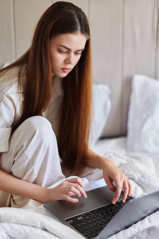 woman laptop bed bedroom pajamas working: Young woman sitting on bed in bedroom wearing pajamas and working on laptop, focused on screen for remote work, study or browsing with cozy home atmosphere