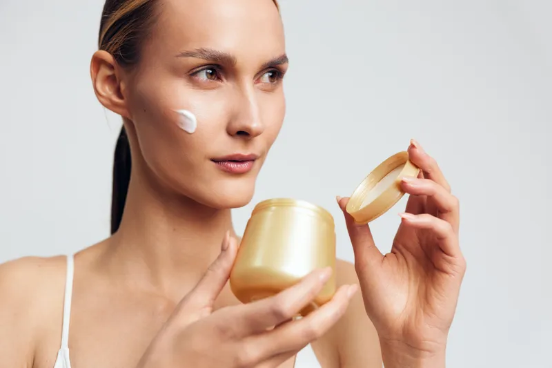 Focused woman applies cream on face holding golden jar with open lid skincare concept isolated on white background closeup portrait beauty routine daily care
