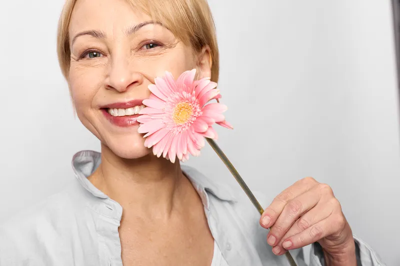 Woman portrait closeup smiling with pink flower held near cheek, soft white background, beauty skincare mood, spring nature, cheerful calm expression.