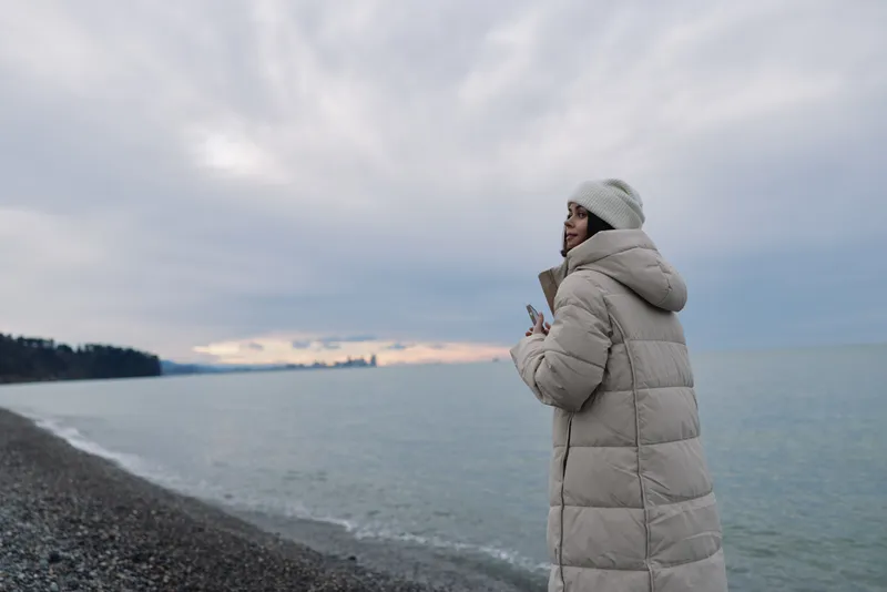 Woman in a long padded coat stands on a pebbled shoreline, gazing at a misty sea under a cloudy sky, winter fashion and coastal calm in a solitary moment