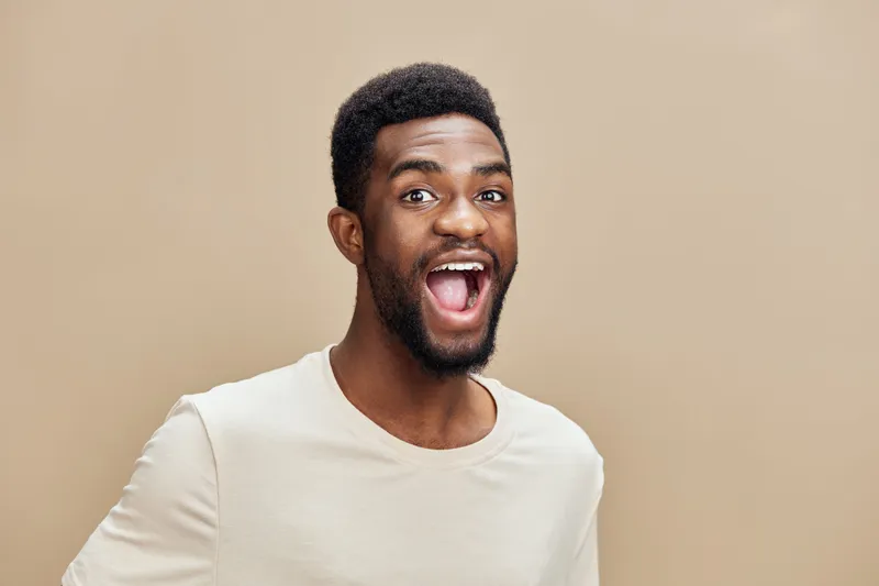 Happy Afro American man smiling widely in a light beige shirt against a neutral background, conveying joy and positivity in lifestyle imagery