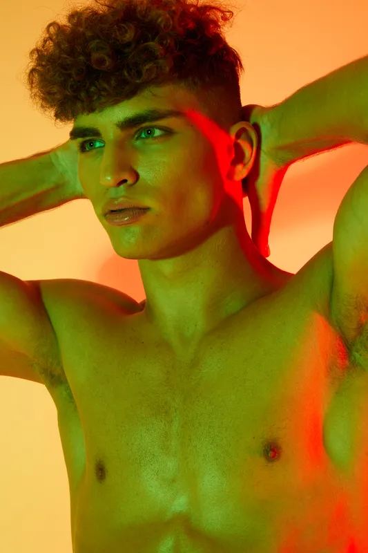 Young man with curly hair posing shirtless with hands behind head, colorful lighting highlighting his face and upper body in studio portrait photography.