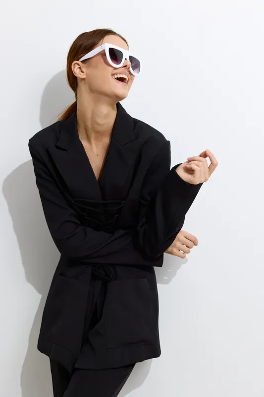 Fashion woman wearing black oversized blazer and white sunglasses laughing joyfully against white background with clean minimalist style and modern vibe.